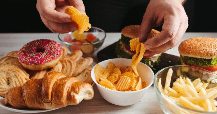 Mesa com alimentos ultraprocessados como batata frita, hambúrguer, donuts, salgadinhos e pães, representando dieta não saudável.