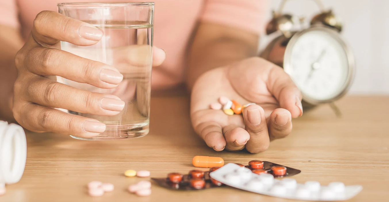  Mão feminina segura copo de água e medicamentos sobre mesa, com relógio ao fundo.