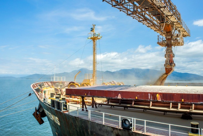 Navio cargueiro sendo carregado com grãos em terminal portuário, com mar e montanhas ao fundo.