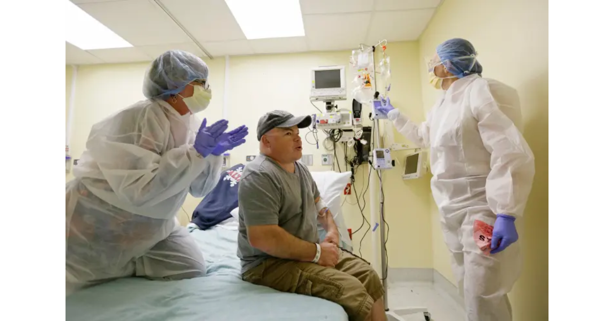 Paciente sentado em leito hospitalar sendo atendido por dois profissionais de saúde com roupas de proteção