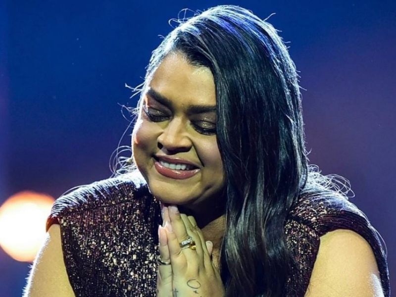 Preta Gil sorrindo com as mãos unidas em gesto de gratidão durante show, iluminada por luz de palco.