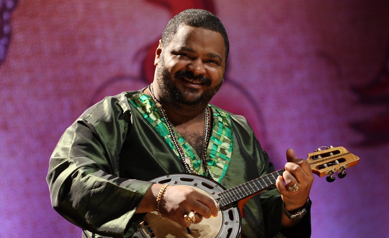 Arlindo Cruz sorrindo e tocando cavaquinho no palco com camisa verde