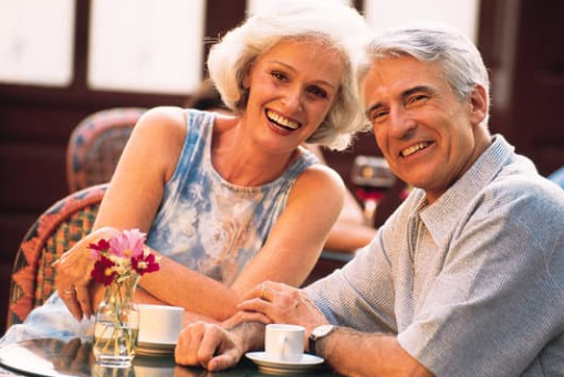 Casal idoso sorridente sentado à mesa com xícaras de café e flores, em ambiente agradável