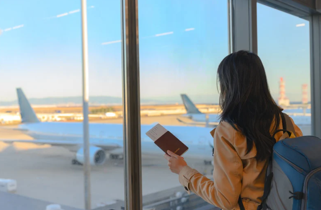 Mulher com passaporte observa avião pela janela de aeroporto.