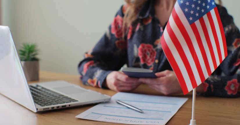 Pessoa segura passaporte ao lado de formulário de visto americano e bandeira dos EUA.