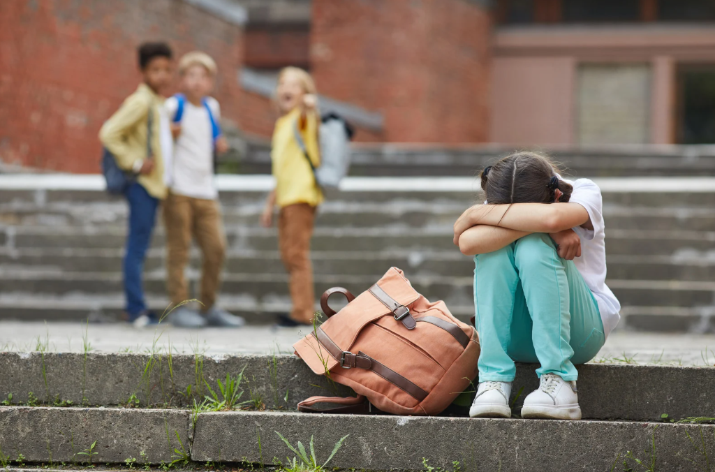 Criança sentada nos degraus da escola com a cabeça baixa, enquanto colegas ao fundo riem e apontam, representando bullying escolar.