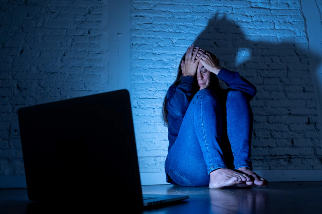 Jovem sentada no chão com expressão de angústia diante de um notebook, simbolizando os efeitos do cyberbullying.
