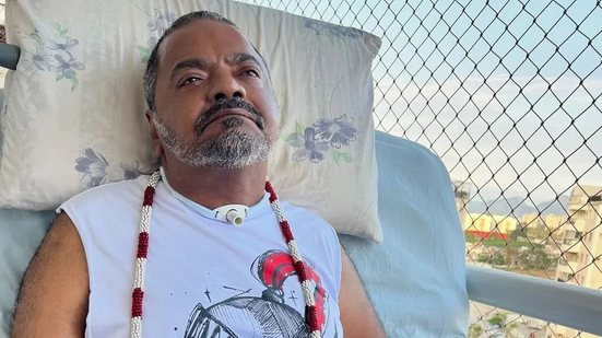 Arlindo Cruz deitado em cadeira hospitalar em casa, usando colar e traqueostomia