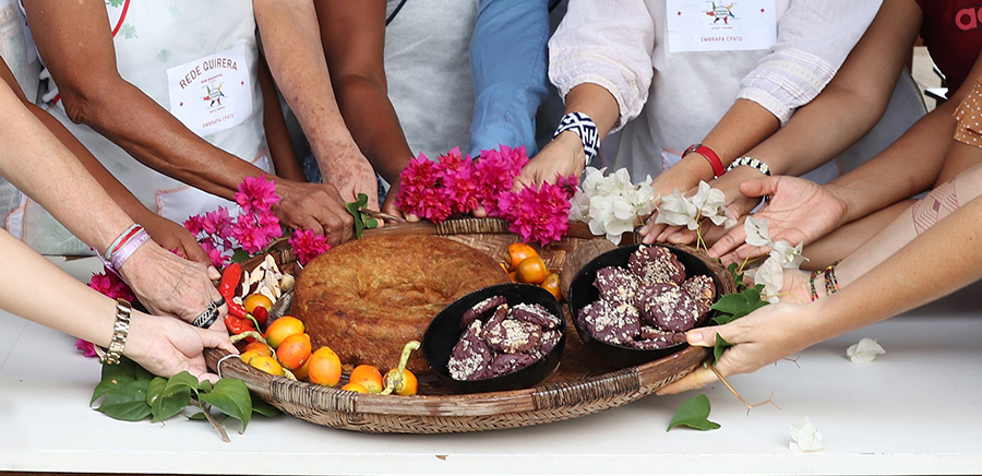 Mãos de mulheres e jovens seguram uma bandeja com alimentos típicos amazônicos, como bolo, farinha e frutos, decorados com flores.