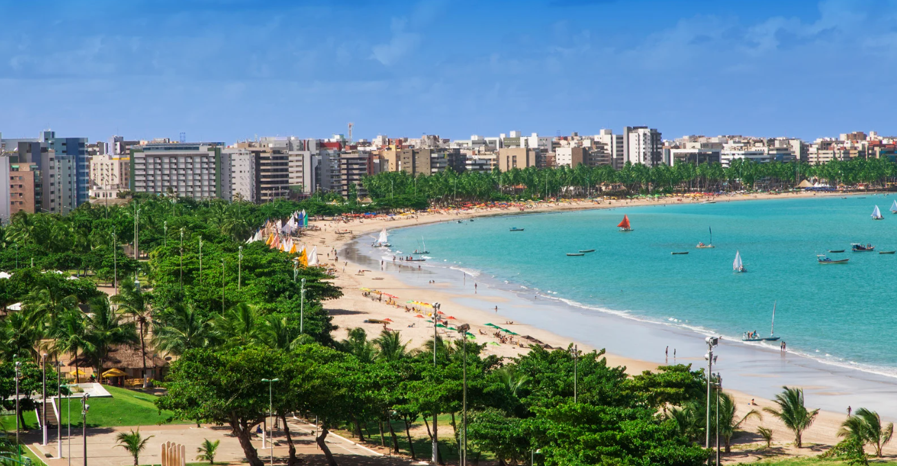 Vista aérea de praia urbana com mar azul, prédios ao fundo, coqueiros e barcos de passeio.