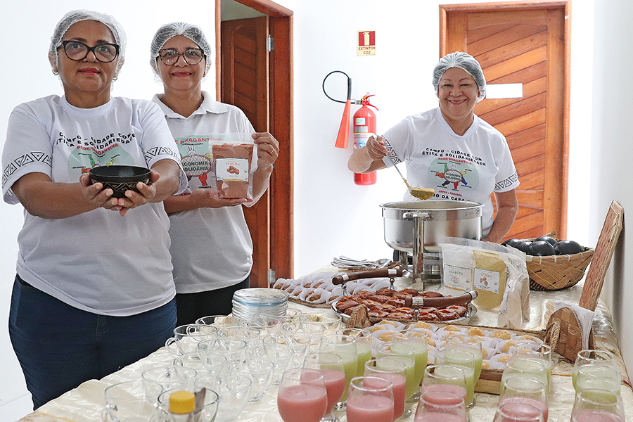 Três mulheres da Rede Bragantina, usando toucas e camisetas padronizadas, exibem produtos de farinha artesanal e preparam alimentos com ingredientes amazônicos.